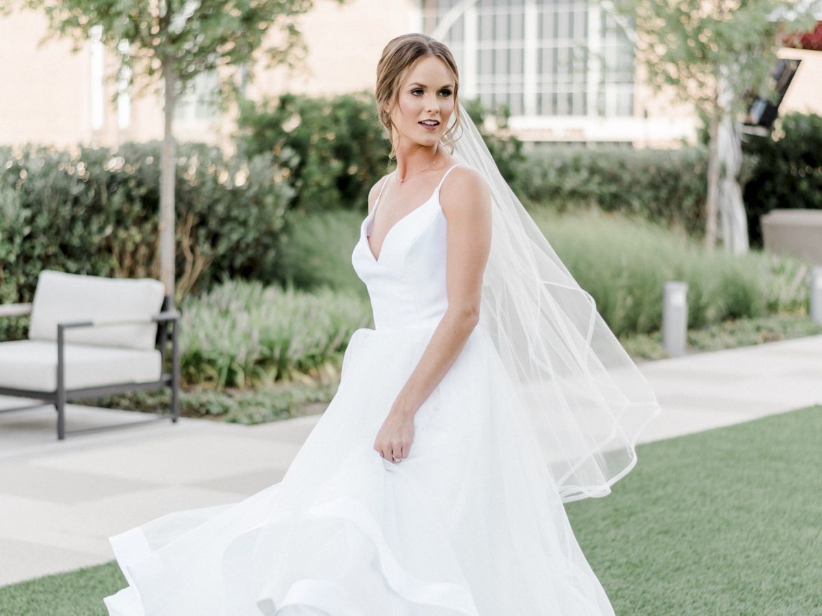 A bride in a white gown and veil poses on a garden lawn outside a bright building, captured mid-walk.