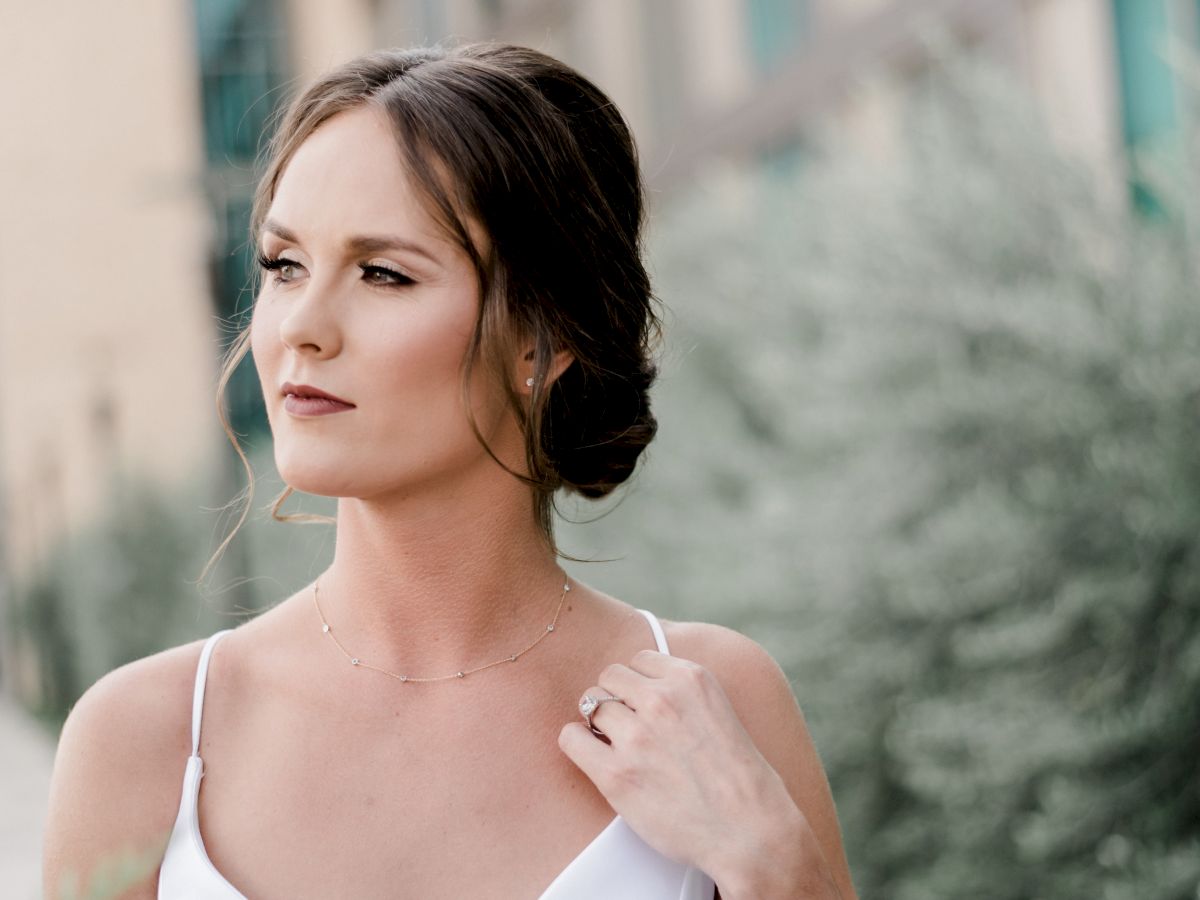 A woman in a white dress poses outdoors, wearing a delicate necklace and ring, with a soft blurred building background behind her.