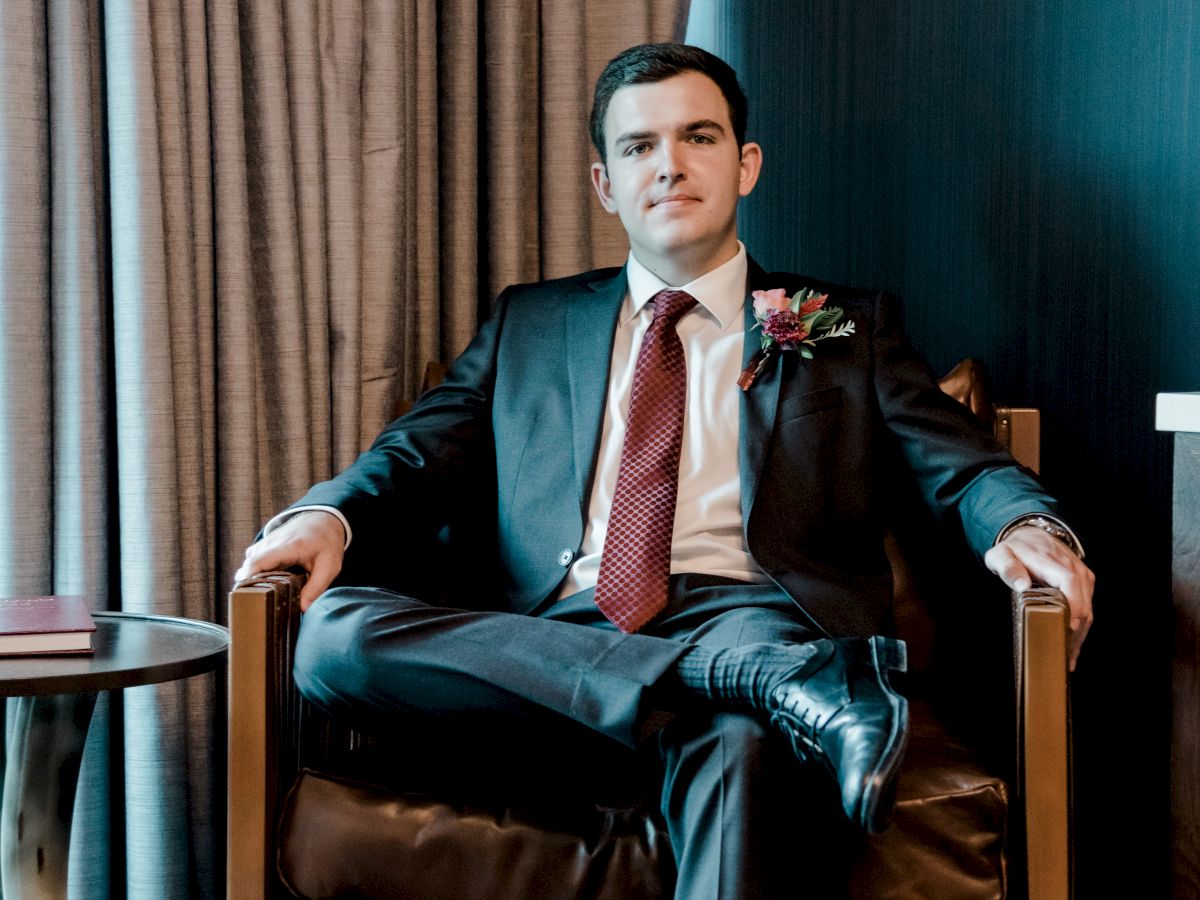 A man in a teal suit with a red tie and boutonniere sits in a leather chair beside curtains in a formal room.