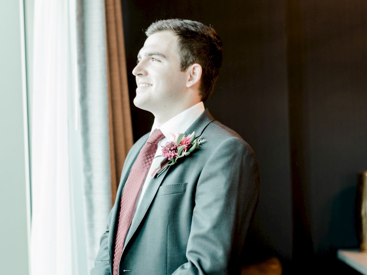 A man in a teal suit and pink tie wears a flower boutonniere, smiling indoors near a window.