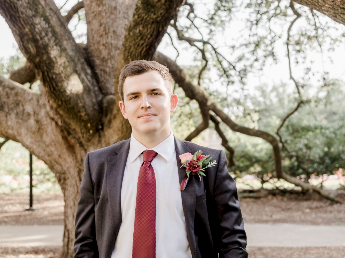 A young man in a dark suit with a red tie and boutonniere stands outdoors, posing beneath large trees.