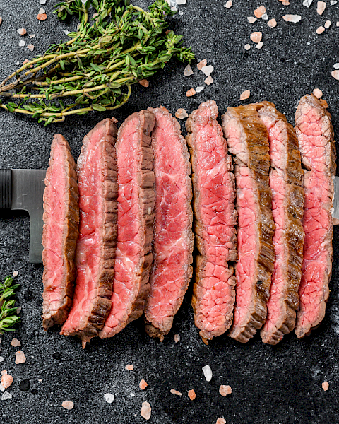 Sliced seasoned beef steak with herbs, salt, and pepper sits on a dark board beside a chef&rsquo;s knife and cherry tomatoes.