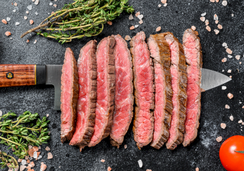 Sliced seasoned beef steak with herbs, salt, and pepper sits on a dark board beside a chef&rsquo;s knife and cherry tomatoes.