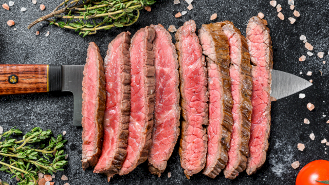 Sliced seasoned beef steak with herbs, salt, and pepper sits on a dark board beside a chef&rsquo;s knife and cherry tomatoes.