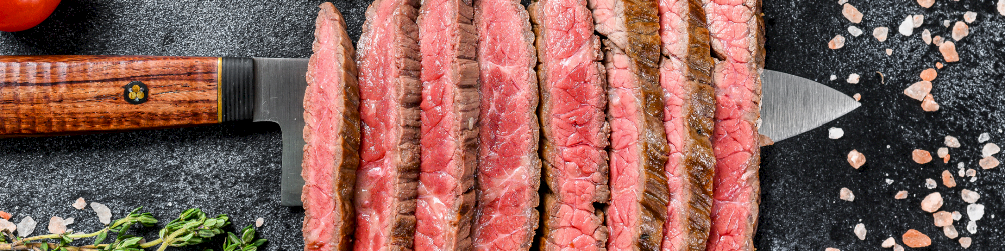 Sliced seared steaks rest on a knife on a dark board, with fresh herbs, pink salt, and tomatoes scattered around.