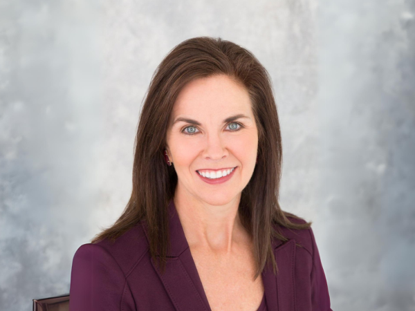 The image shows a woman with brown hair, wearing a purple blazer, smiling at the camera against a grey background.