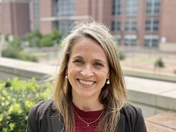 A smiling woman stands outdoors in front of a brick university-style building and greenery, on a cloudy day.