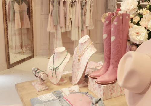 A boutique storefront display with pastel dresses, jewelry, cowboy boots, and flowers in a vintage-themed shop.