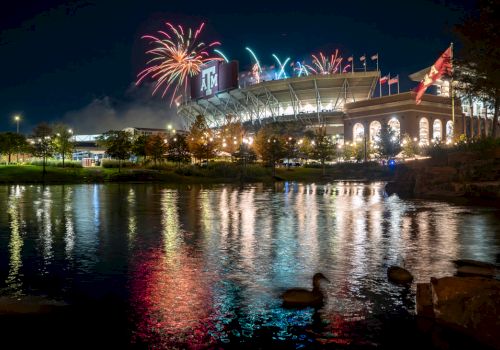 Fireworks burst over a lit stadium and flags, reflected in a calm lake at night.