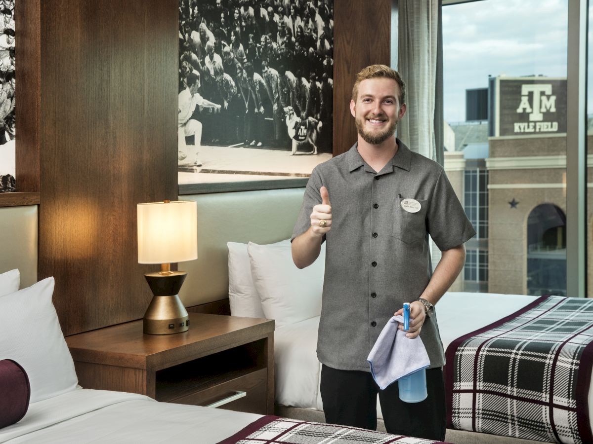 A hotel staffer smiles and gives a thumbs-up beside a bed, holding a towel in a guest room.