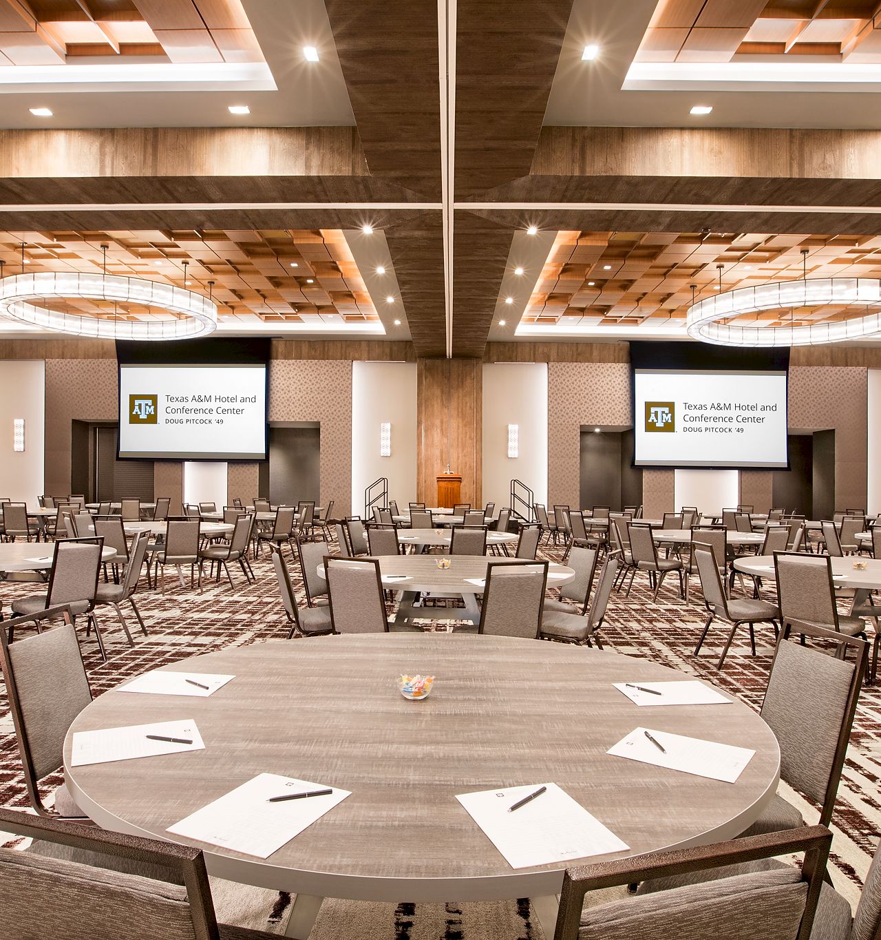 A spacious modern conference room set up for meetings, with round tables, chairs, overhead lights, and Texas A&M presentation screens.