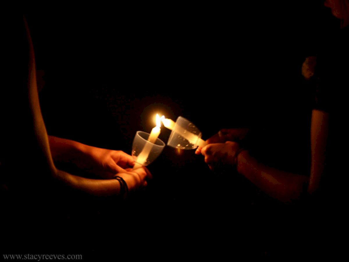 Three hands are holding candles with plastic covers in the dark, sharing the flame from one candle to another.