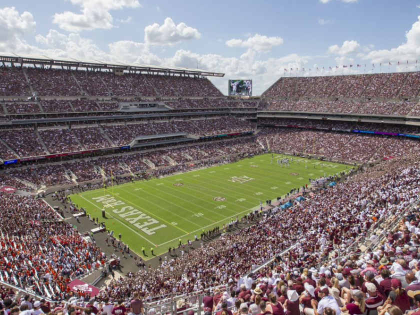 A packed football stadium with fans filling the stands as teams play on the green field under a cloudy sky.