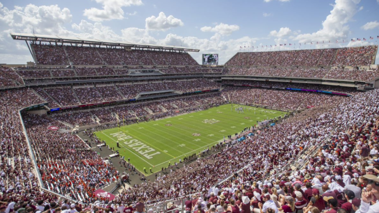 A packed football stadium with fans filling the stands as teams play on the green field under a cloudy sky.