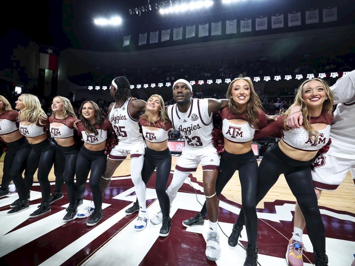 A group of cheerleaders and basketball players are standing on a court with arms linked, smiling and celebrating under bright lights.