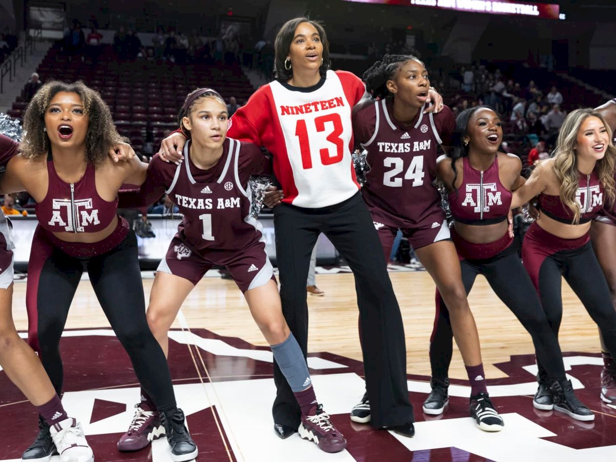 A group of women wearing Texas A&M outfits and one in a "NINETEEN 13" shirt are standing together on a basketball court, looking enthusiastic.
