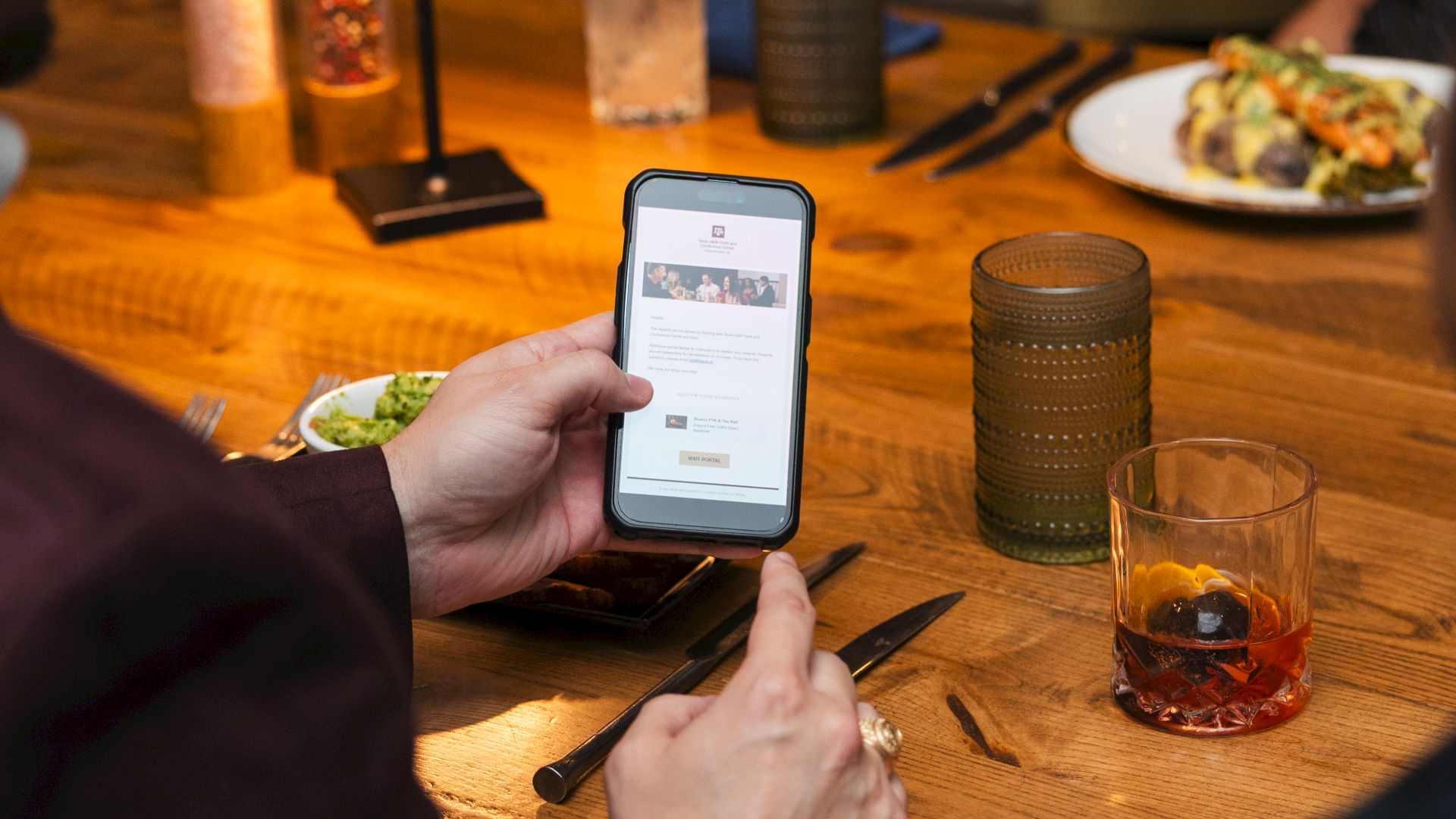 A person at a restaurant holds a smartphone, seemingly checking a menu or ticket, while drinks and food sit on the table.