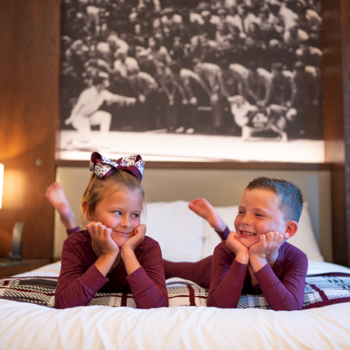 Two kids on a bed, smiling and posing with their hands by their cheeks, with one wearing a colorful bow headband.