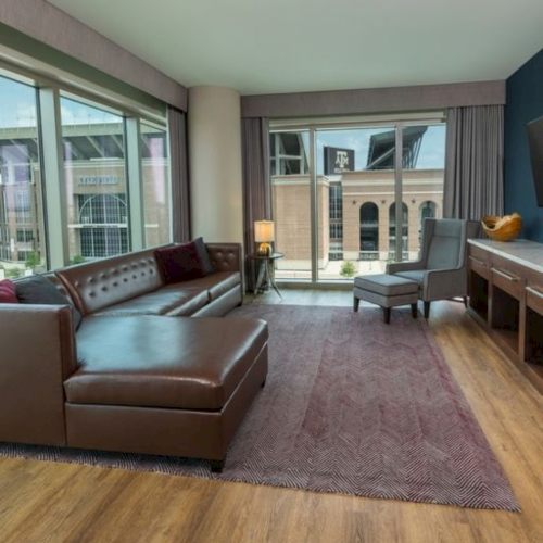 A modern living room with brown leather sofa, armchair, rug, coffee table, and large sliding windows overlooking a campus building.