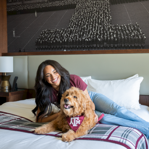 A woman smiles on a bed with her golden doodle wearing a Texas A&M bandana in a cozy hotel room.