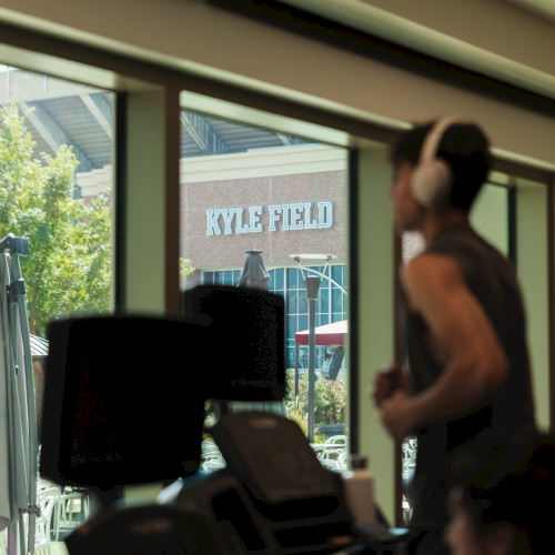 A blurred spectator with headphones watches from inside as &ldquo;Kyle Field&rdquo; shows through the window.