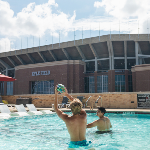 A boy throws a splashy beach ball in a pool outside Kyle Field stadium on a sunny day.