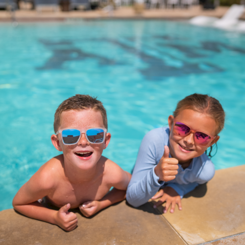 Two kids in a sunny pool wearing goggles, smiling and giving thumbs-up from the pool edge.