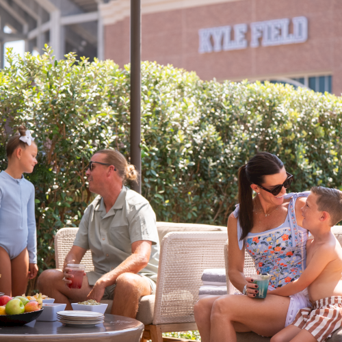 A family enjoys a sunny outdoor picnic, sharing drinks and food on a patio with trees and a school sign behind.