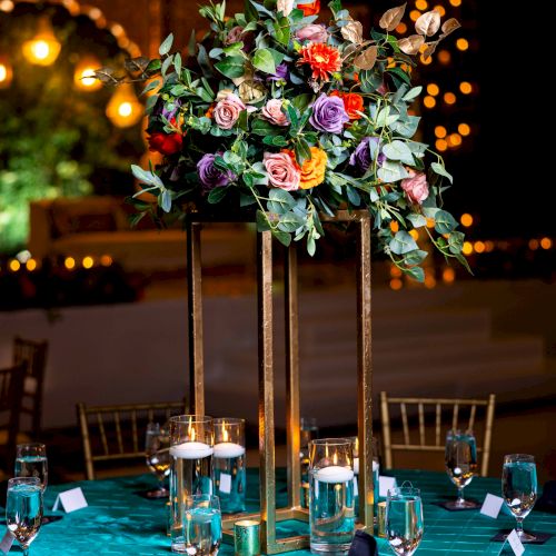 Colorful rose-and-greenery centerpiece on a banquet table, surrounded by glowing candles and warm bokeh lights.