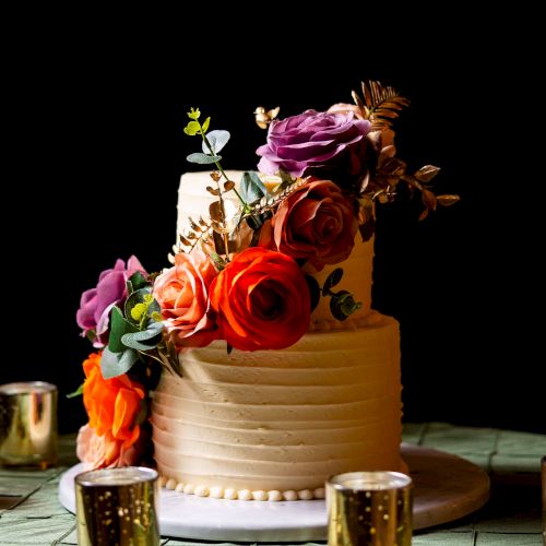 A tiered cake topped with colorful roses sits on a table with gold cups, glowing against a dark background.