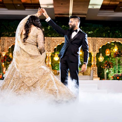A groom in a black tux lifts a bride in a sparkling gown at a lit wedding stage, with smoke and flowers behind them.