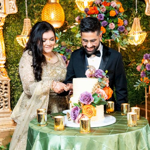 A couple in formal wear cuts a decorated wedding cake amid gold lights and colorful flowers.