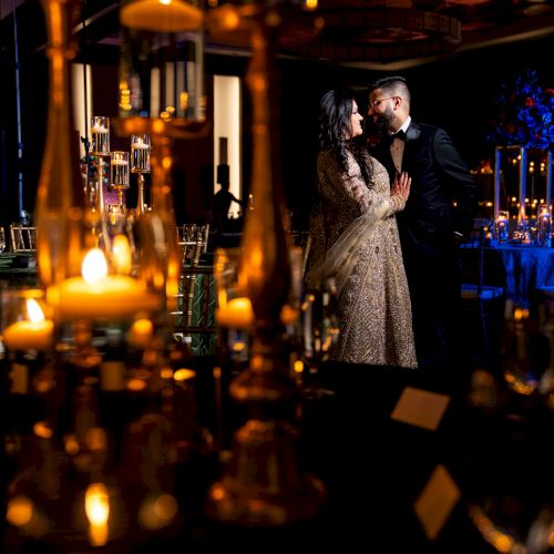 A bride and groom in elegant attire share a close moment at a candlelit, warmly lit wedding reception.
