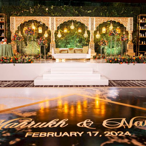 An ornate event stage with a gold floral arch, glowing chandeliers, lush bouquets, and a lit white couch centerpiece.