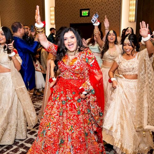 A group of women in festive traditional outfits dance with joy at an indoor celebration, waving hands and smiling.