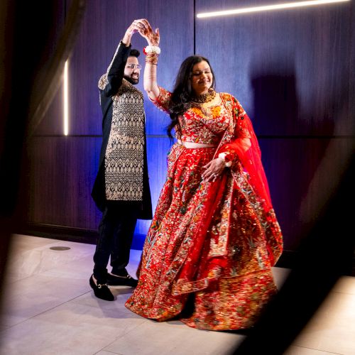 A couple dances in traditional red-and-gold attire at an event, with raised hands under blue stage lights.