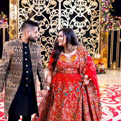 A man and woman in festive wedding attire stand together indoors amid floral d&eacute;cor and an ornate gold backdrop.
