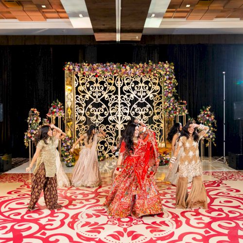 A wedding stage with ornate floral decor and a group of women in elegant dresses dancing in front of seated guests.