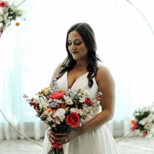 A woman in a white dress holds a colorful wedding bouquet under a curved arch at an indoor ceremony.