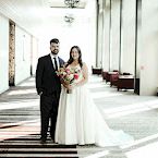 A newlywed couple stands together in a hallway, the groom in a black suit and the bride in a white gown holding flowers.