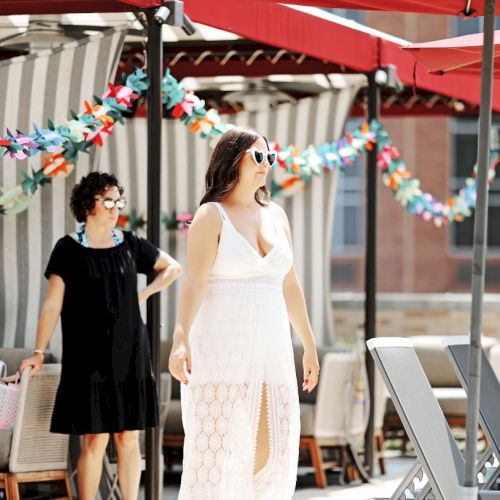 Two women stroll at a sunny outdoor patio under red tents, surrounded by colorful floral garlands and deck chairs.