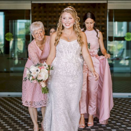 A bride in a white lace dress walks down steps with an older woman, holding a bouquet, while bridesmaids follow behind her.