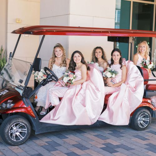 A group of women in pink dresses sits in a red golf cart outside, holding bouquets and smiling like a wedding party.