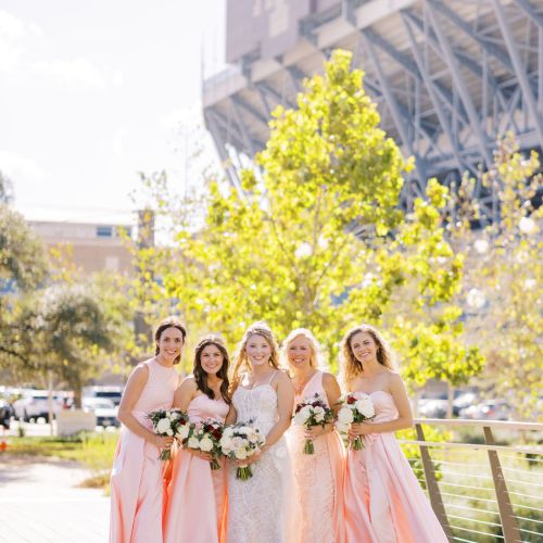 Five bridesmaids in pastel dresses pose with bouquets on a sunny deck beneath a stadium facade.