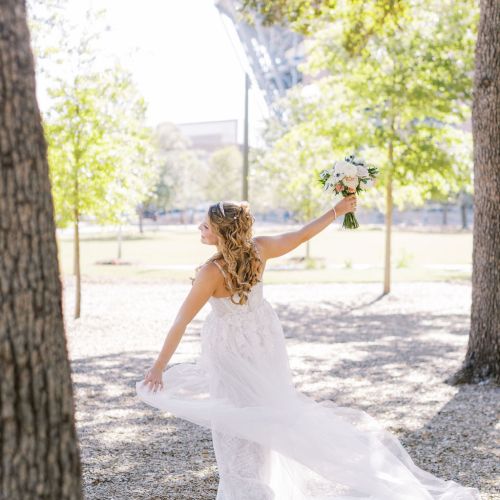 A bride in a white dress sits among trees holding a bouquet in the sunny park.
