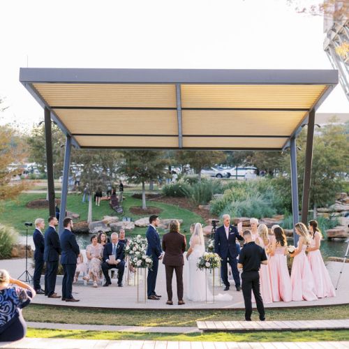 An outdoor wedding ceremony on a stage beneath a pavilion, with guests and a bridal party around flower-covered arches.