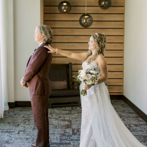 A bride in a lace gown holds a bouquet while an older man adjusts her jacket in a hotel hallway.