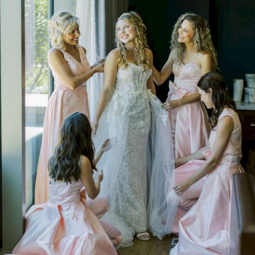 Three women in pastel gowns help a bride in a bright room before a ceremony, while others bustle around her.