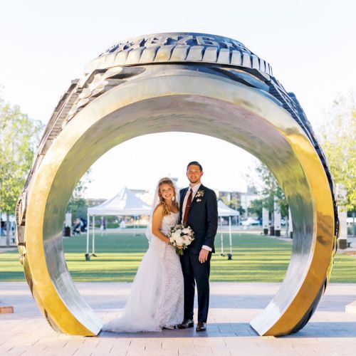 A bride and groom pose for a wedding photo inside a large metallic ring sculpture in a park courtyard.