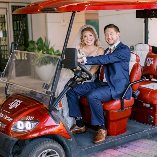 A smiling bride and groom pose in a red golf cart outside a building, ready for their big day.